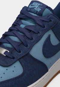 Κοντινή λήψη ενός μπλε παπουτσιού Nike Air Force 1 με σκουρόμπλε κορδόνια, υφασμένα πάνελ και σόλα από καουτσούκ χρώματος καφέ.