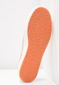 Podeszwa sneakersa z fakturowaną pomarańczową gumową podeszwą zewnętrzną i białą wkładką środkową, ustawiona pod kątem na białym tle ściany i podłogi.