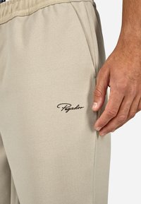 Bézs színű, puha anyagból készült melegítőnadrág, oldalsó zsebekkel; a combon hímzett barna "Pegador" logóval.