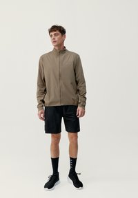 Lätt, beige zip-up jacka med hög krage, i kombination med svarta shorts. Har minimal varumärkesprofil och en slät textur. Svarta sportskor.