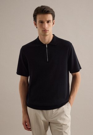 Joven hombre vestido con una camiseta tipo polo negra de manga corta con cremallera frontal y pantalones beige, de pie contra un fondo claro y liso.