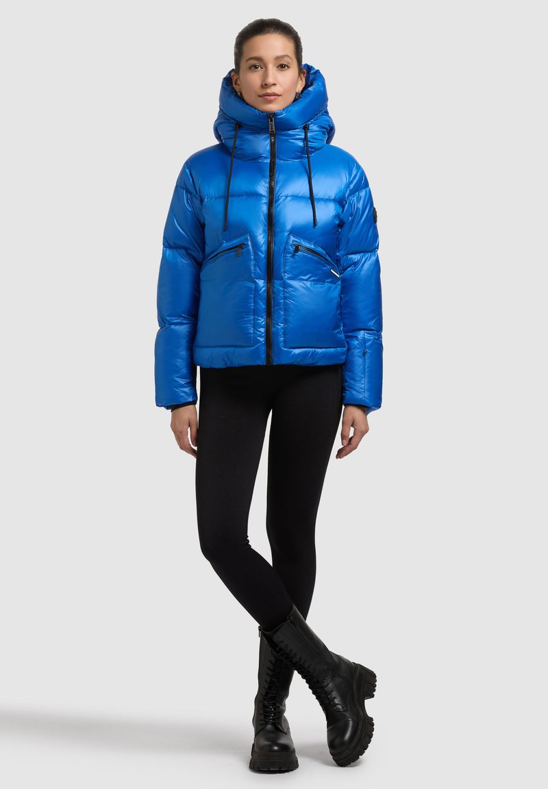 Modra puhovka z visokim ovratnikom in vezalkami, s dvema žepoma na prsih z zadrgo, kombinirana s črnimi legicami in masivnimi črnimi škornji.