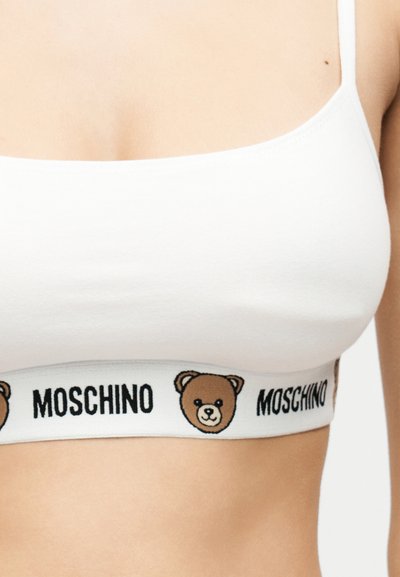 Λευκό αθλητικό σουτιέν με καφέ πρόσωπα αρκουδάκια και το κείμενο «MOSCHINO» στη λαστιχένια ταινία κάτω από το στήθος, φορεμένο από ένα άτομο.