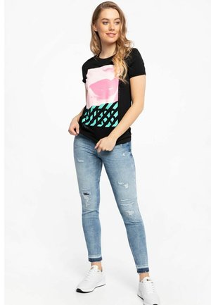 T-shirt nera con una grafica di labbra rosa e un motivo geometrico teal. Indossato con jeans blu strappati e sneakers bianche. Texture del tessuto liscia.