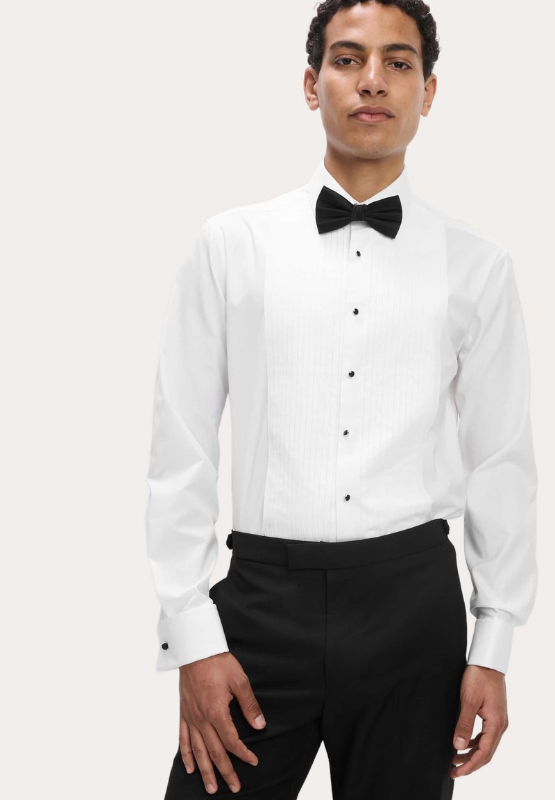 Weißes Hemd mit schwarzer Fliege. Mit plissierter Vorderseite, schwarzen Knöpfen und französischen Manschetten mit schwarzen Akzenten. Getragen mit schwarzer Hose.