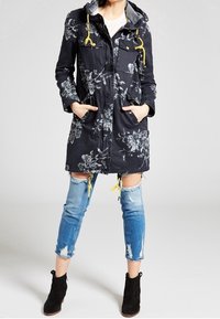 Tmavě modrý park s květinovým vzorem a kapucí, žlutými šňůrkami a čtyřmi předními kapsami. Nosený přes roztrhané modré džíny a černé kotníkové boty.