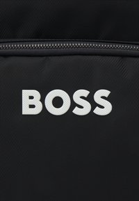 Tessuto nero con una texture diagonale che presenta un logo in gomma bianca ben visibile con la scritta "BOSS," situato sopra una chiusura con zip.