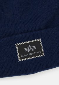 Námornícka pletená čiapka so štvorcovým čiernym štítkom s bielym prešívaním a textom loga "ALPHA INDUSTRIES." Mäkčená textúra, ohnutý dizajn.