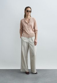 Cardigan rosa claro con botones y cuello redondeado, combinado con pantalones anchos crema y zapatos planos marrones. La modelo también lleva gafas de sol oscuras.