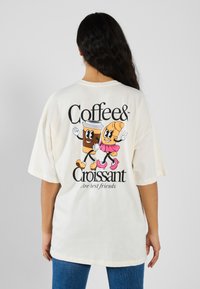 Ljus beige oversized t-shirt med tecknade bilder av en leende kaffekopp och en croissant, svart text som säger "Kaffe & Croissant."