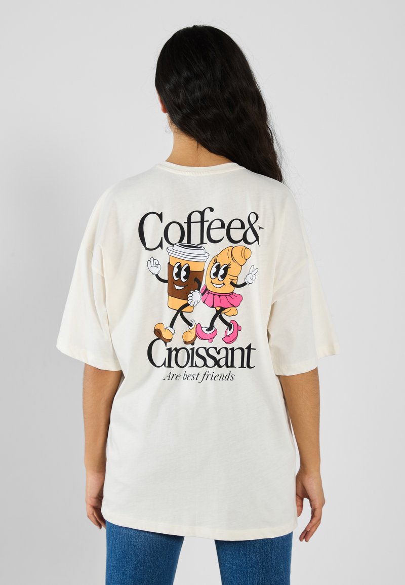 Ljus beige oversized t-shirt med tecknade bilder av en leende kaffekopp och en croissant, svart text som säger "Kaffe & Croissant."