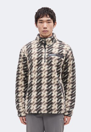 Young man wearing a beige, black, and brown patterned fleece pullover with a half-zip and chest pocket, standing against a plain background.