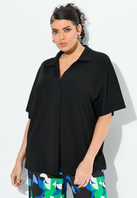 Černá košile s krátkým rukávem a límcem, s volným střihem a detailním skladem na přední části. Nosené s barevnými vzorovanými kalhotami.