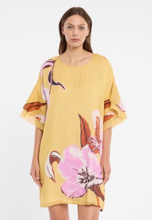 Frau trägt ein locker sitzendes gelbes Kleid mit großen pinken und braunen Blumenprints, steht vor einem schlichten weißen Hintergrund.