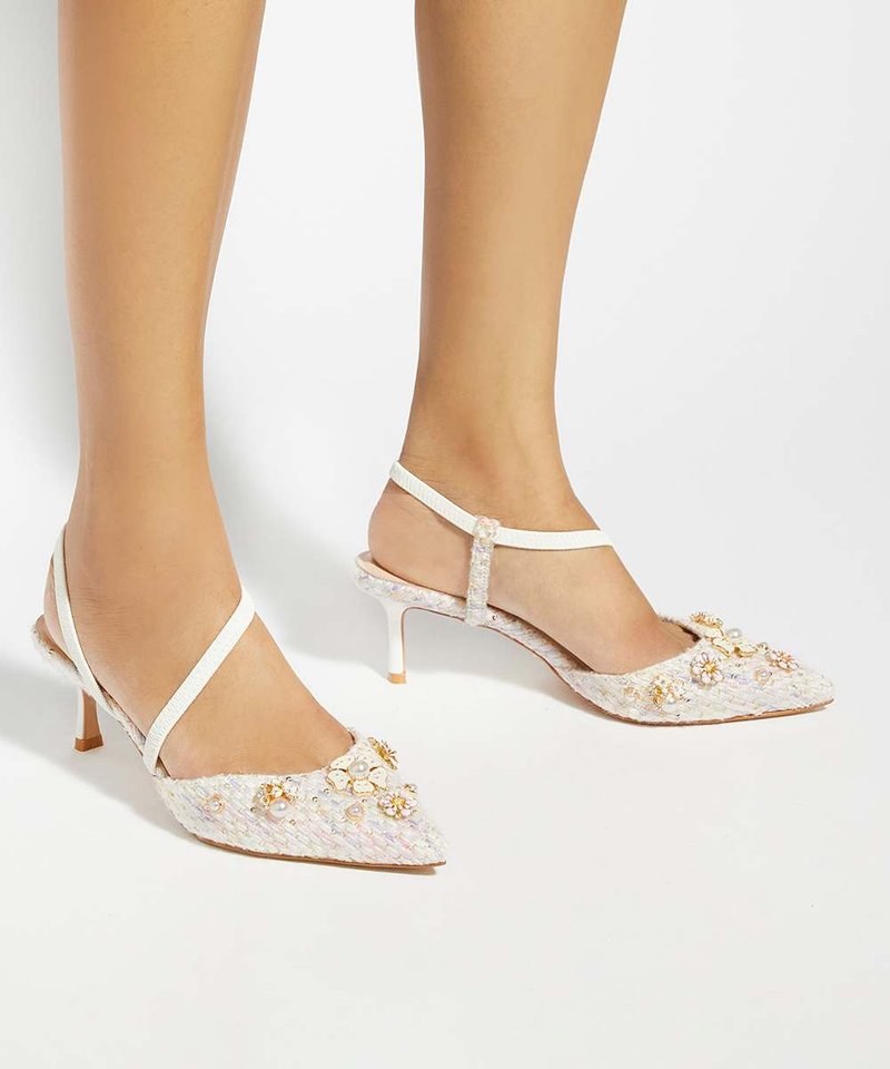 Pieds portant des talons slingback blancs à bout pointu décorés de perles et d’ornements floraux sur un fond clair.