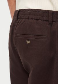 Κοντινή λήψη ατόμου που φοράει σκούρα καφέ παντελόνια με ελαστική μέση και κουμπωτή πίσω τσέπη, συνδυασμένα με λευκό ριμπ τοπ.