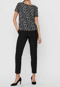 Donna che indossa una blusa a maniche corte con motivo bianco e nero, pantaloni neri slim-fit e scarpe con tacco nero, in piedi con le mani nelle tasche.