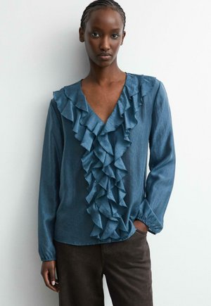 Woman wearing a blue long-sleeve blouse with layered ruffles down the front and dark pants, standing against a white wall.