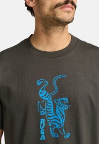 Donkergrijze katoenen T-shirt met een blauwe afbeelding van een gestileerde tijger en de merknaam "RVCA" in het wit. Ronde hals met korte mouwen.