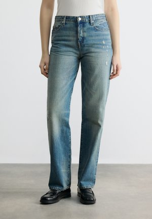 Γυναίκα που φοράει τζιν μπλε χρώματος με υψηλή μέση και ίσια φάρδια, με ελαφριά φθορά στο δεξί μηρό, συνδυασμένα με μαύρα μοκασίνια σε γκρι πάτωμα.