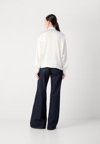 Blouse blanche à manches longues avec des poignets, assortie à un jean large bleu foncé. La blouse a un léger éclat ; le jean présente des coutures classiques.