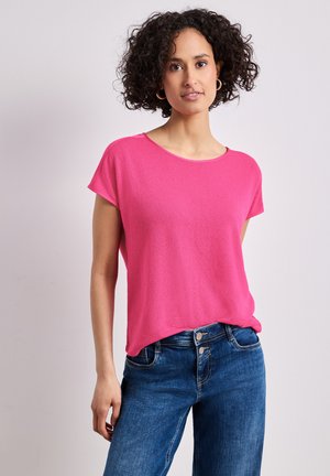 Kvinde med krøllet hår iført en lys pink kortærmet top og blå jeans med høj talje, står mod en ensartet lys baggrund.