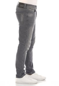 Vaqueros de mezclilla gris con corte ajustado, diseño de cinco bolsillos y un ligero desgaste. Presenta una etiqueta negra en la cintura y se combina con zapatillas blancas.