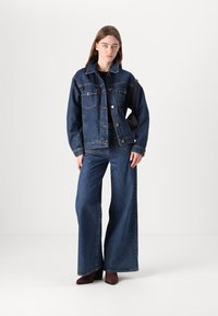 Jeansjacka med framfickor och knappar i mörkblått, kombinerad med vida mörkblå jeans, båda med synliga sömnadsdetaljer.