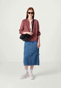 Světle růžová pletená bomber bunda, bílá košile, džínová sukně s kapsami, černá crossbody taška, bílé tenisky a černé sluneční brýle.