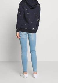 Felpa con cappuccio blu navy decorata con motivi di libellule dorate, abbinata a jeans skinny blu chiaro e sneaker bianche. Presenta un grande cappuccio e polsini a coste.
