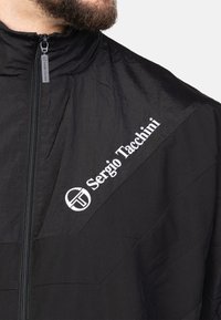 Schwarze Zip-Jacke aus leichtem Material mit strukturiertem Design. Weißes besticktes Logo auf der Brust und ein hoher Kragen.