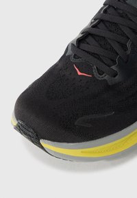 Adidași negri de alergare cu un exterior din plasă, având un accent roșu, detalii gri și o talpă galbenă. Vârful are o formă rotunjită cu elemente texturate.
