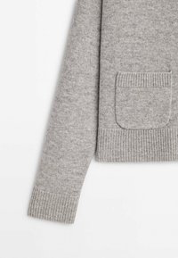 Manche et partie avant d'un pull gris clair en tricot avec des bordures côtelées aux poignets et à l'ourlet, ainsi qu'une petite poche appliquée sur le bas devant.