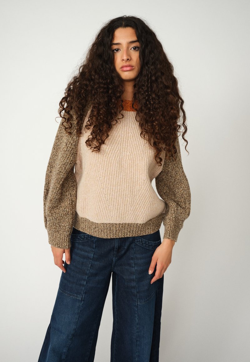 Rauschiger Strickpullover mit einem beigen Körper und braunen strukturierten Ärmeln, ausgestattet mit einem gerippten Saum und weiter Silhouette. Kombiniert mit dunkelblauen Jeans.