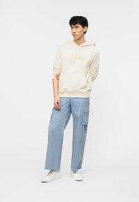 Νέος άνδρας που στέκεται, φοράει κρεμ φούτερ με κουκούλα και λογότυπο, ανοιχτό μπλε παντελόνι cargo και λευκά αθλητικά παπούτσια, μπροστά σε ένα απλό λευκό φόντο.