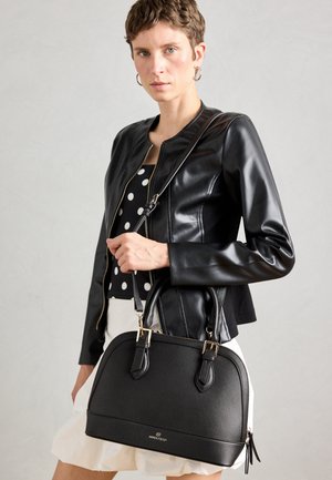Woman wearing black leather jacket and polka dot top holds black Anna Field handbag with gold hardware against light background.