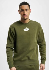 Olivinės spalvos Nike džemperis su apvaliu kaklu, gumuotais rankogaliais ir liemeniu. Priekyje yra baltas Nike logotipo užrašas. Minkšta, tekstūruota medžiaga.