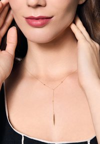 Złoty naszyjnik z długim, smukłym wisiorkiem, połączony z małymi koralikami na delikatnym łańcuszku, noszony na nagiej szyi.