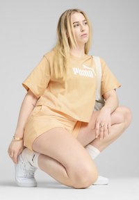 Ferskenfarvet bomulds-t-shirt med hvid Puma-logo, matchende shorts og hvide sneakers. Model knæler med synlige accessories.