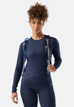 Mujer con cabello rizado que lleva una camiseta ajustada de manga larga azul marino y leggings, llevando una mochila negra con un tubo de hidratación azul.
