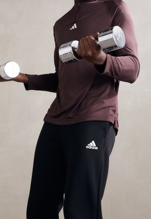 Persoon gekleed in een bordeauxrode top met lange mouwen en zwarte Adidas-broek, tilt twee chromen dumbbells van 3 kg tegen een effen achtergrond.