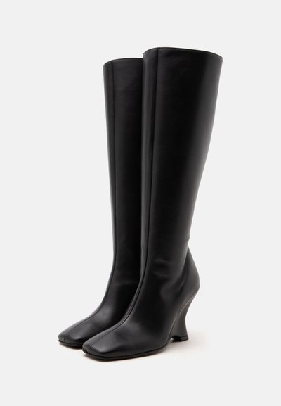 Bottes hautes en cuir noir avec talon compensé et bout carré, debout sur un fond blanc uni.