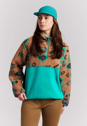 Femme portant une casquette bleu sarcelle et un pull polaire marron et bleu sarcelle à motifs avec un pantalon beige, debout devant un fond clair uni.