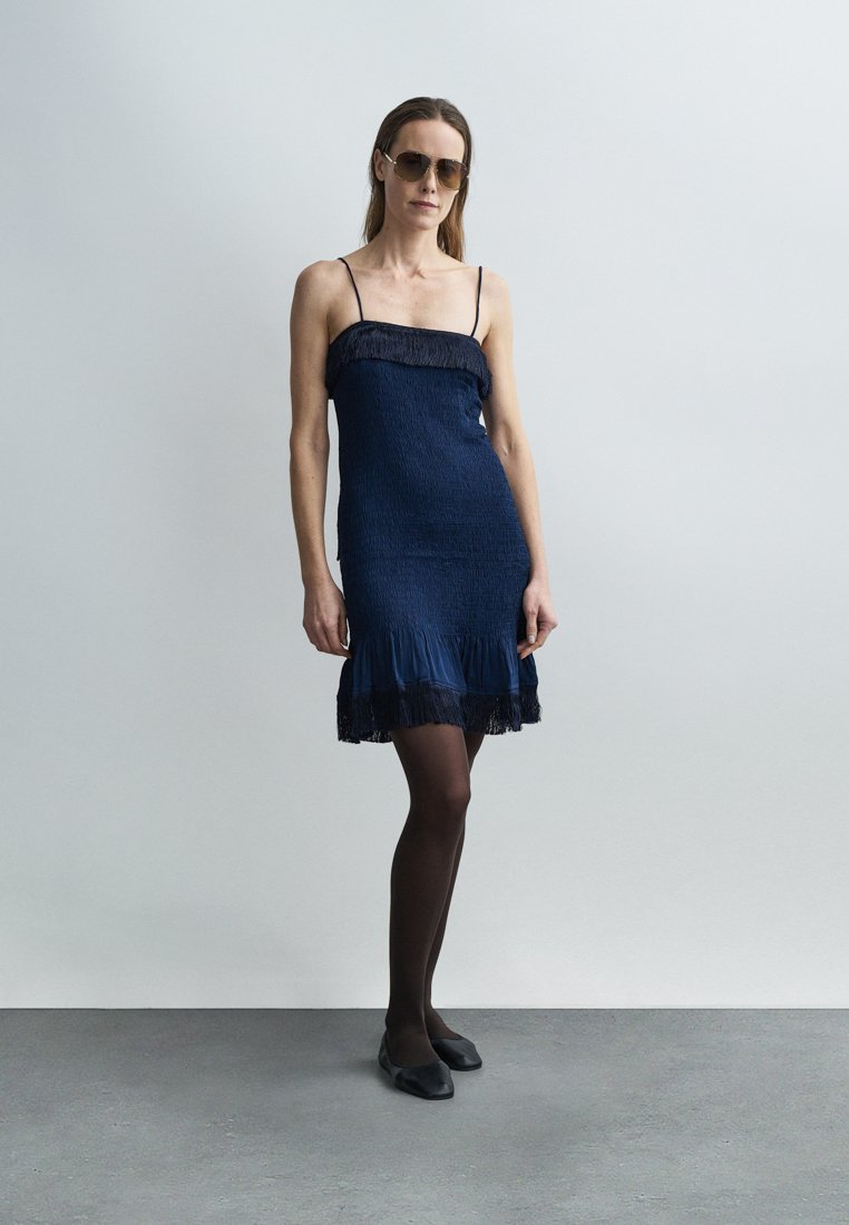 Robe bustier bleu marine en tissu texturé avec ourlet à volants. Portée avec des collants noirs et des chaussures plates ; modèle portant des lunettes de soleil.