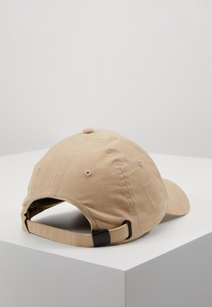 Beżowa bawełniana czapka z daszkiem z regulowanym paskiem z tkaniny i czarną metalową klamrą, leżąca na białej powierzchni.