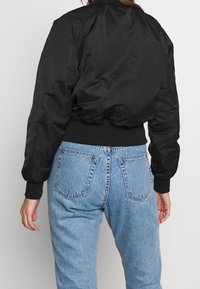 Czarna kurtka bomber z gładką powierzchnią, ściągaczami przy mankietach i w pasie; noszona na tle niebieskich dżinsów z wysokim stanem, z tylnymi kieszeniami.