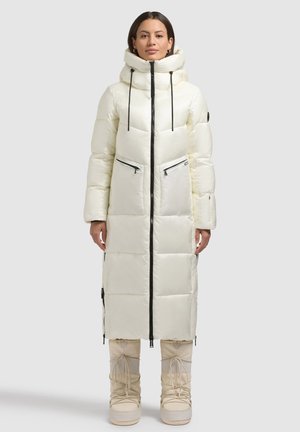 Long, padded white coat with a high collar, front zipper, and two zippered pockets. Paired with light beige knee-high boots.