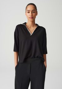 Schwarzes Poloshirt mit kurzen Ärmeln und V-Ausschnitt, verziert mit weißen Paspelierungen am Kragen. Glattes Material, lässige Passform.