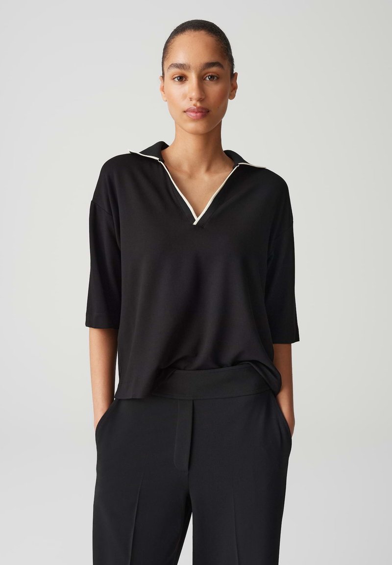 Schwarzes Poloshirt mit kurzen Ärmeln und V-Ausschnitt, verziert mit weißen Paspelierungen am Kragen. Glattes Material, lässige Passform.