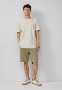 Beige kurzärmliges Hemd aus strukturiertem Stoff, kombiniert mit olivgrünen Shorts. Das Modell trägt weiße und beige Sneakers mit braunen Akzenten.